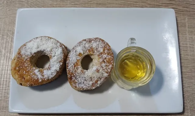 Two ring shaped pastries dusted with sugar and a small glass of golden liquid, on a white plate