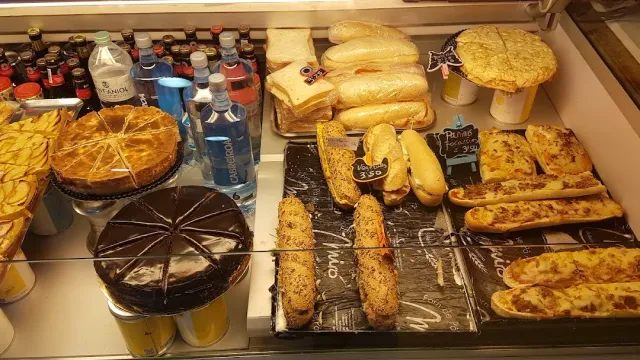 A display case with sandwiches, baked baguettes, whole cakes and drink bottles behind