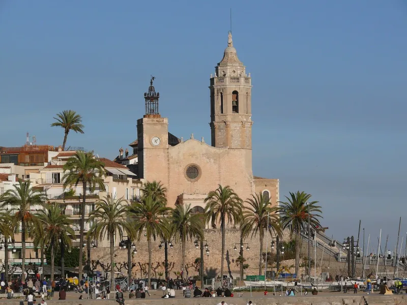 A stone church with a bell tower on a cliff above the sea, ringed by palms and white houses