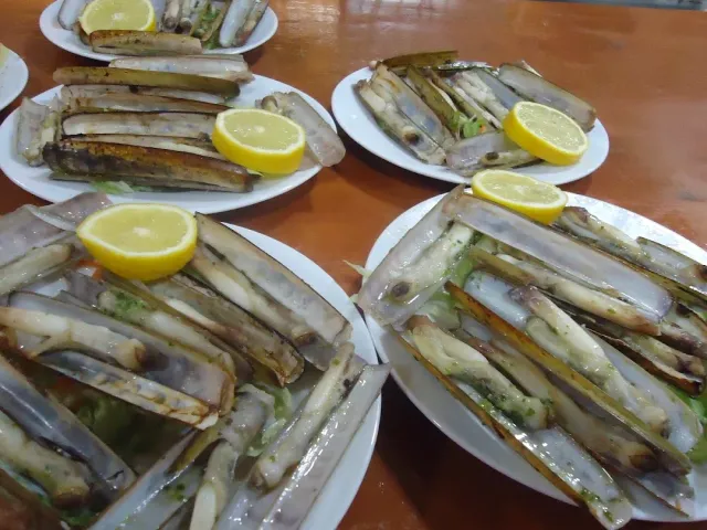 Four plates with grilled razor clams and lemon wedges, on a wooden table