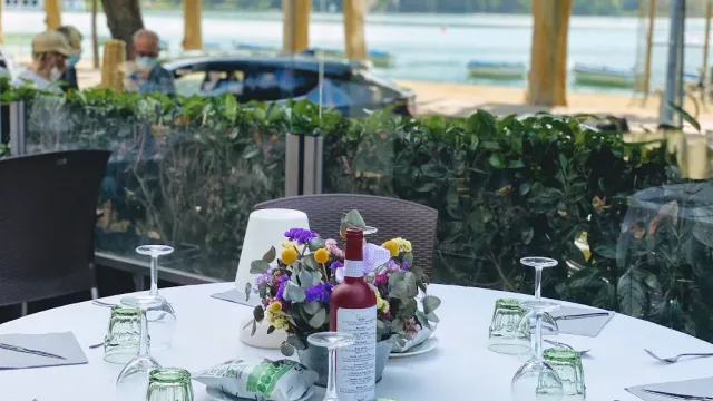 A set round table with a bouquet and a bottle, with a lake and boats behind
