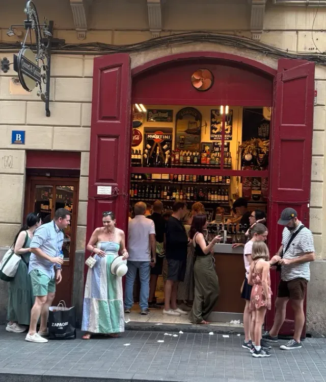 A shop opening in a red frame with shelves of bottles, and customers standing outside