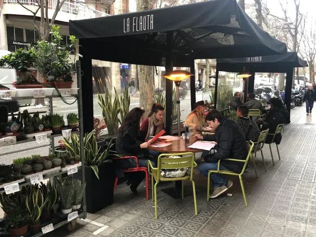 A street terrace under a black canopy with green chairs, diners and a plant shop alongside