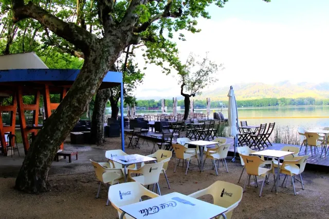White tables and chairs on a lake shore, under large plane trees