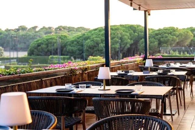 A covered terrace with dark tables, table lamps and flowers facing a pine wood