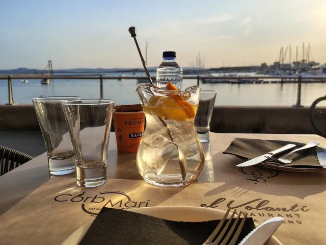 A glass jug with ice and fruit slices on a table, with a boat marina in the background