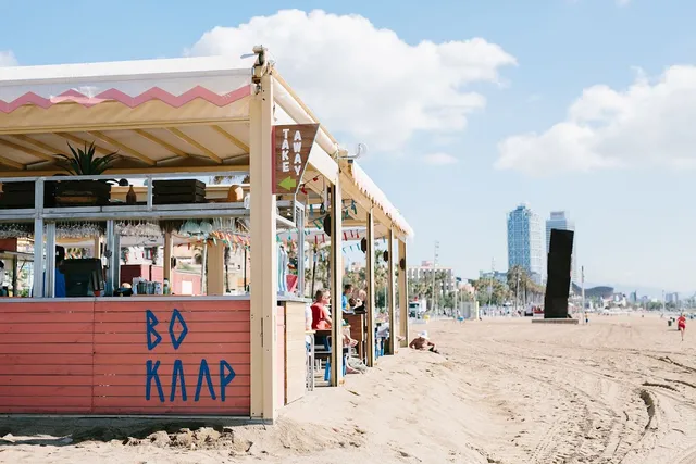 A wooden stall with a striped roof on the sand, with tall towers in the background under a blue sky