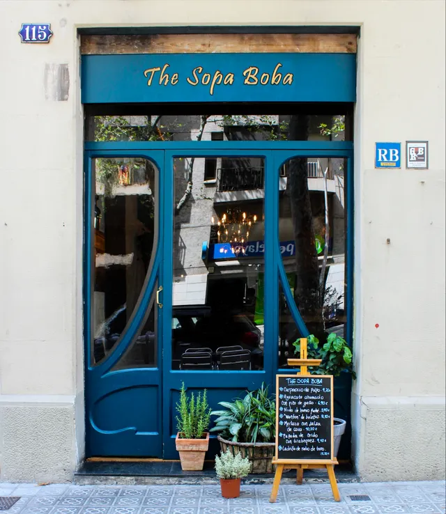 A curved blue entrance door with a gilded sign, potted plants and a chalkboard on an easel