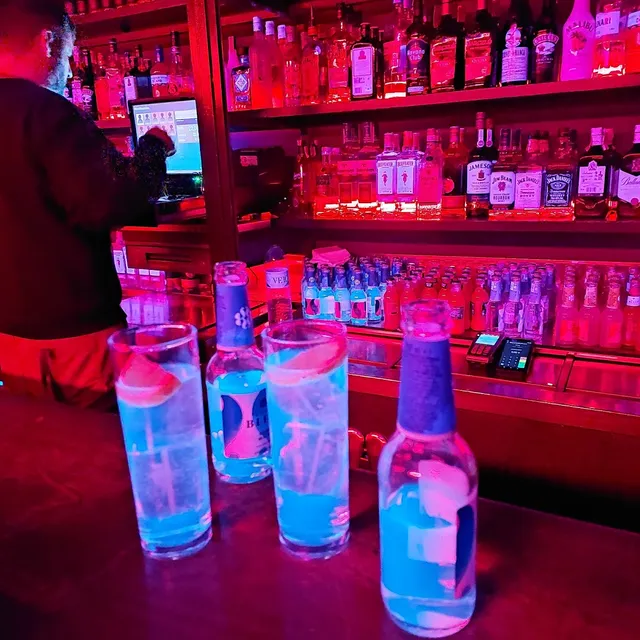 Blue drinks in tall glasses on a bar, facing bottle shelves in red light