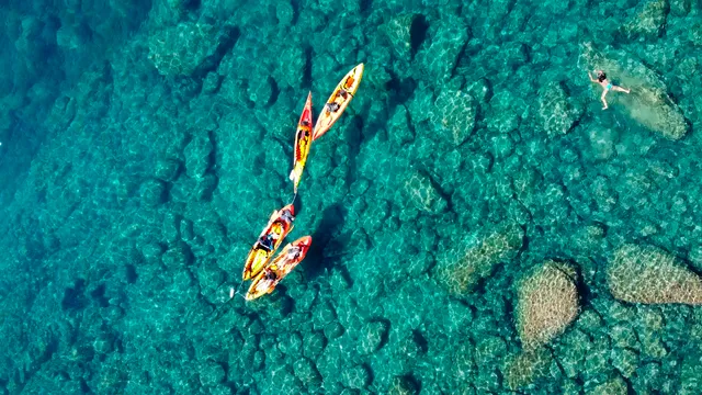 Colorful kayaks and a swimmer in clear turquoise water over rocks, seen from above