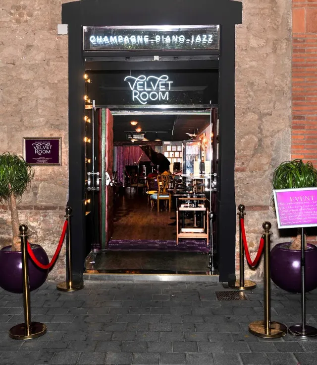 A neon lit entrance in a stone wall, with red ropes and a grand piano inside