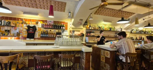 A drinks counter with shelves of bottles, upturned glasses and people seated on stools