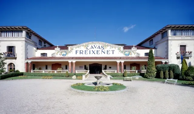 A wide white building with a painted gable, a round fountain and a gravel garden in front