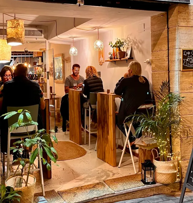 A small space lit in the evening, people seated at tall wooden tables among potted plants