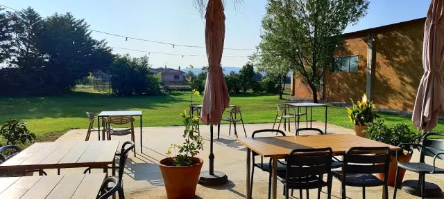 A paved terrace with wooden tables and closed parasols, a lawn and hills behind