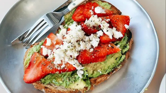 A slice of toast with avocado, strawberries and crumbled white cheese on a metal plate