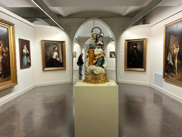 A display hall with framed paintings on white walls and a painted statue on a plinth
