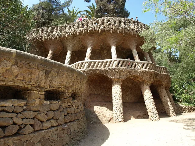 Slanted rough stone columns carrying an upper walkway with a stone balustrade, and a shaded arcade of arches below