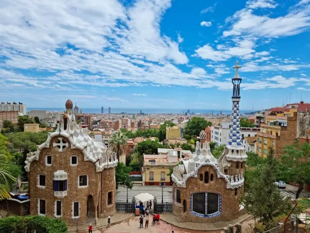 Two stone pavilions with wavy mosaic roofs, with the city and the sea behind them