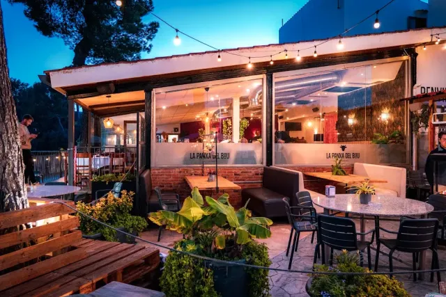 A terrace lit by strings of bulbs with marble tables and plants, at twilight