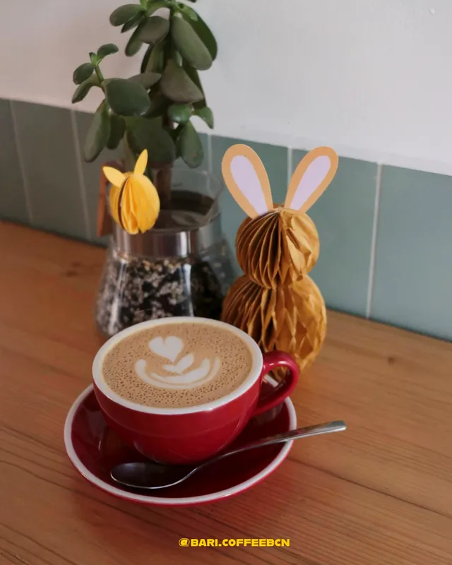 A red cup with latte art, beside a potted plant and a paper rabbit on a wooden table