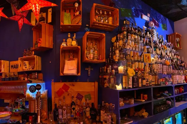 A bar with blue walls, wooden crates and figurines, shelves loaded with bottles and paper stars