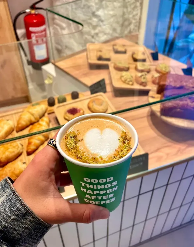 A hand holding a green cup with heart shaped foam, with a pastry case behind it