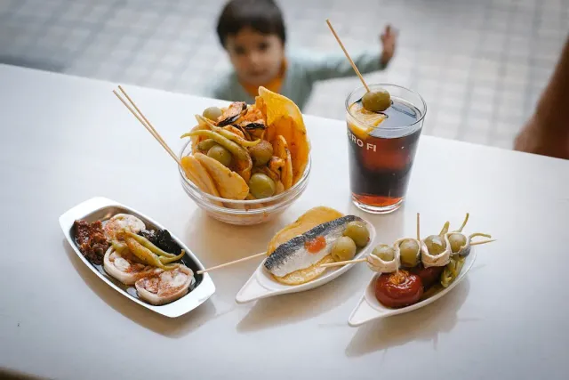 Four small dishes with olives and preserved fish, beside a glass of dark drink