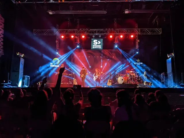 A band performs on a stage lit with blue beams in front of an audience in the dark