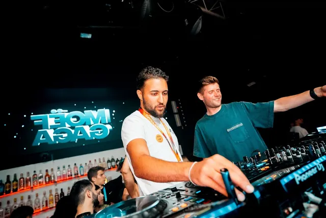 Two DJs at a mixing desk, with a lit screen and a bottle shelf in the background