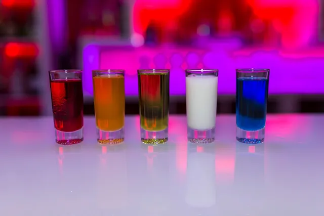 Five colourful shot glasses in a row on a white bar, in pink and purple light
