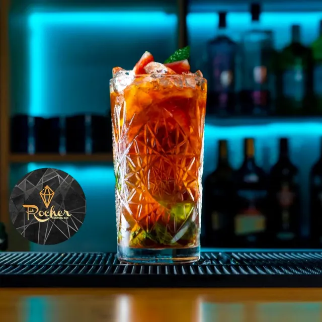 A tall cocktail glass with ice and a red fruit garnish, on a counter in cyan light
