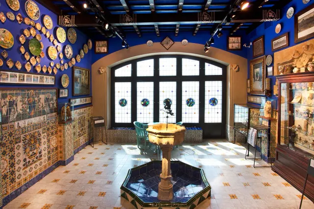 A room with blue walls hung with ceramic plates, a colored glass window and an octagonal fountain