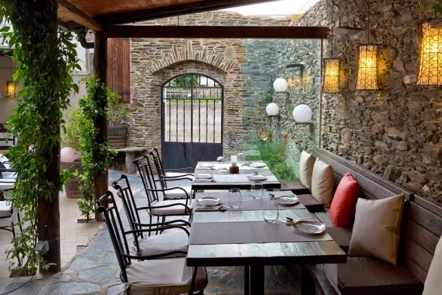 A covered courtyard with wooden tables, a cushioned bench and a stone wall with an iron gate