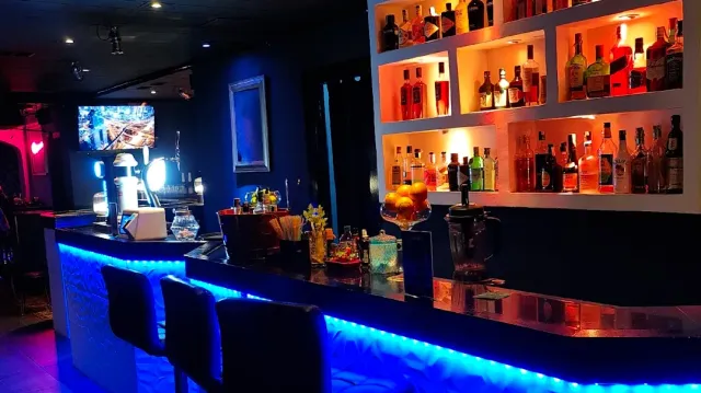 A bar counter in blue lighting, bottle shelves lit in orange and upholstered stools beside it