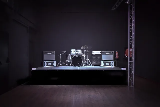 A low stage with a drum kit and amplifiers in a dark, empty hall