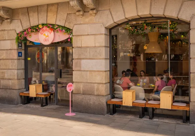 A stone facade with a flower decorated sign, wooden benches and people seated behind a large window