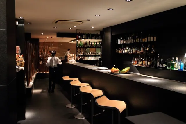 A long dark counter with pale stools and lit bottle shelves along it